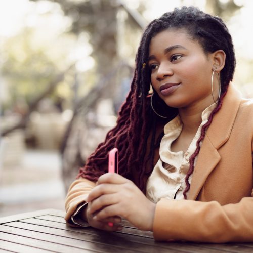 Young african american woman sitting in street cafe and holding smartphone in her hands, close up