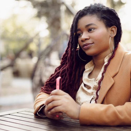 Young african american woman sitting in street cafe and holding smartphone in her hands, close up
